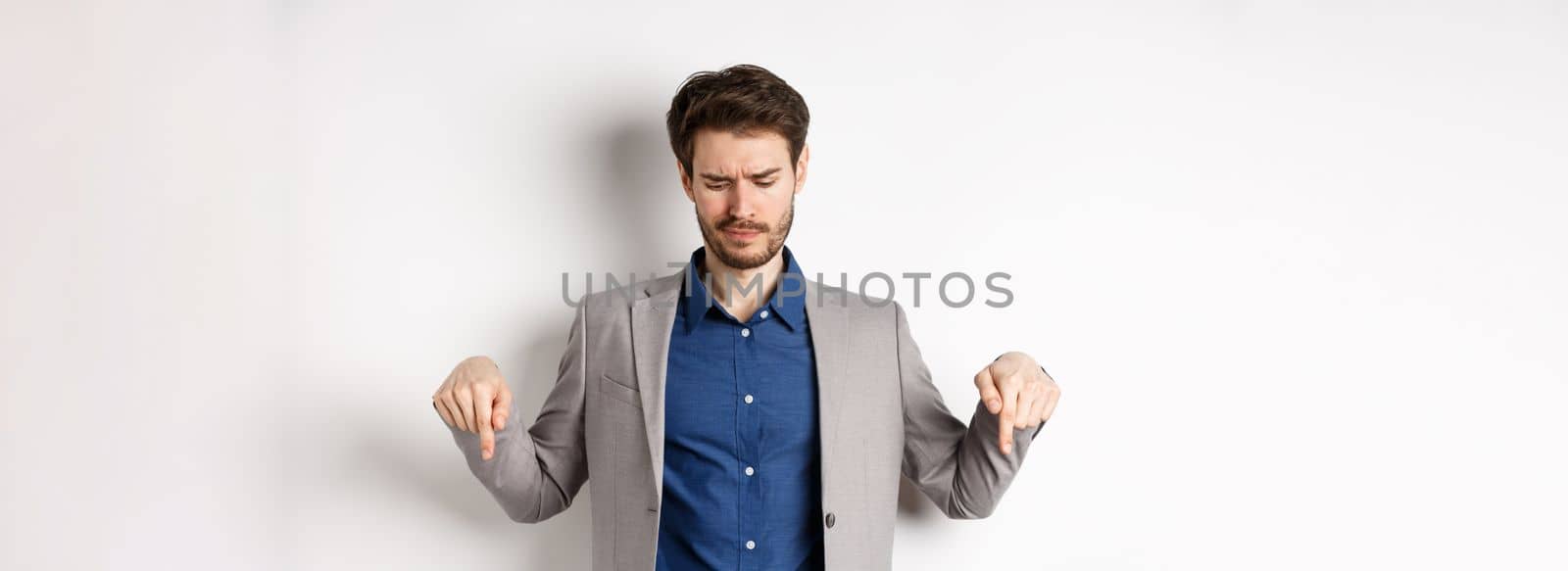 Unimpressed young businessman frowning disappointed, pointing and looking down at bad promotion, standing displeased on white background.