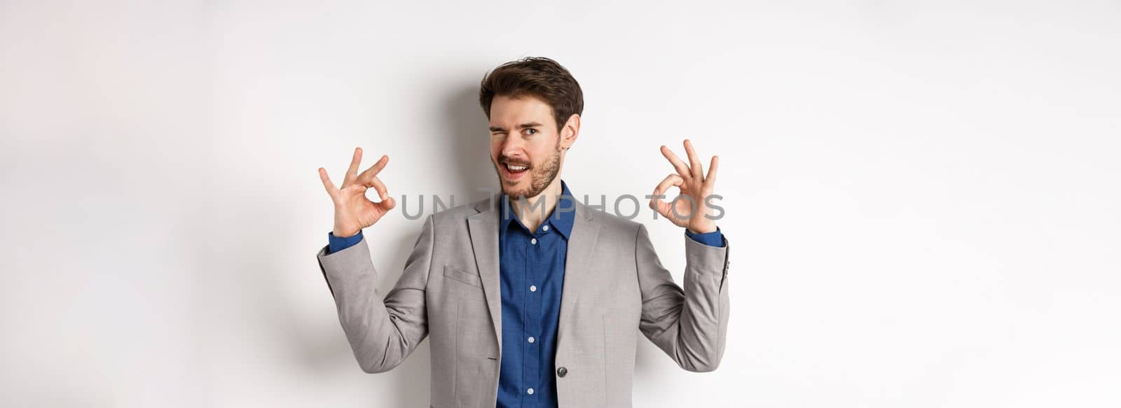Cheeky handsome man in suit winking at you, showing okay signs, all under control gesture, approve good choice, assuring everything OK, standing on white background by Benzoix