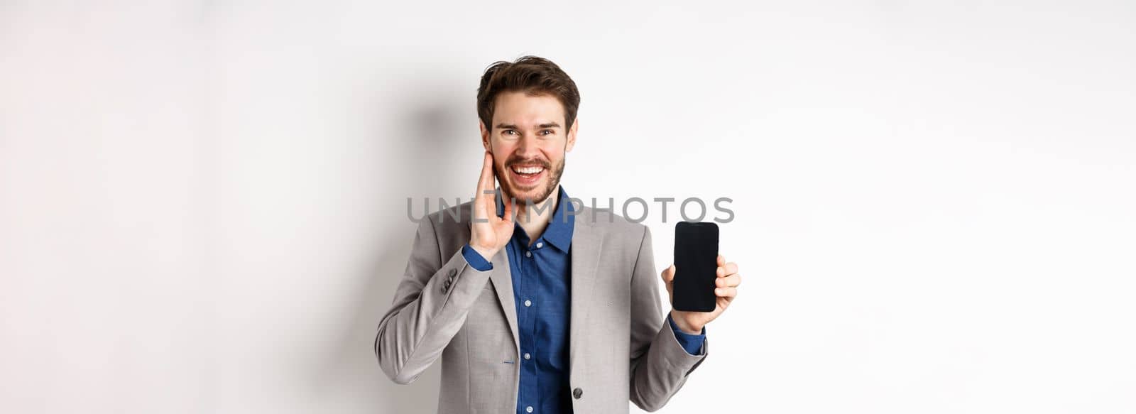 E-commerce and online shopping concept. Happy bearded guy in suit showing empty smartphone screen and laughing, standing on white background.