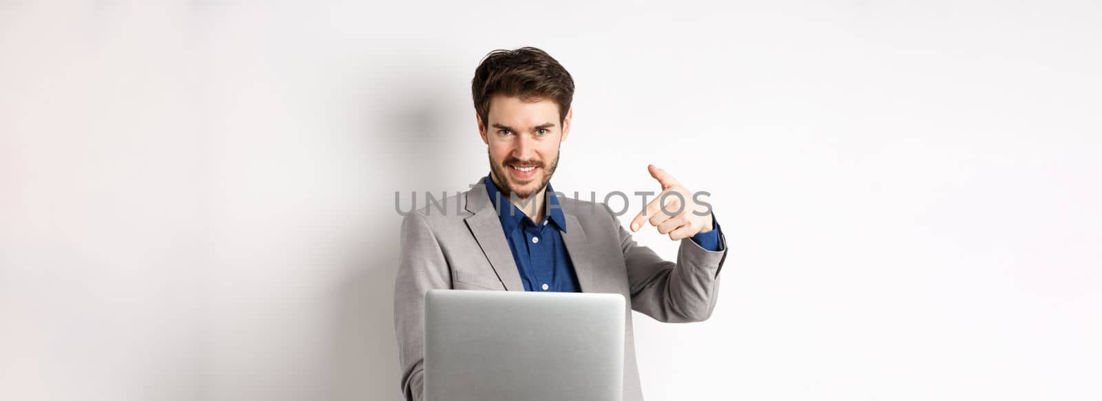 Confident young male entrepreneur working online with laptop, pointing at computer and smiling, standing on white background.