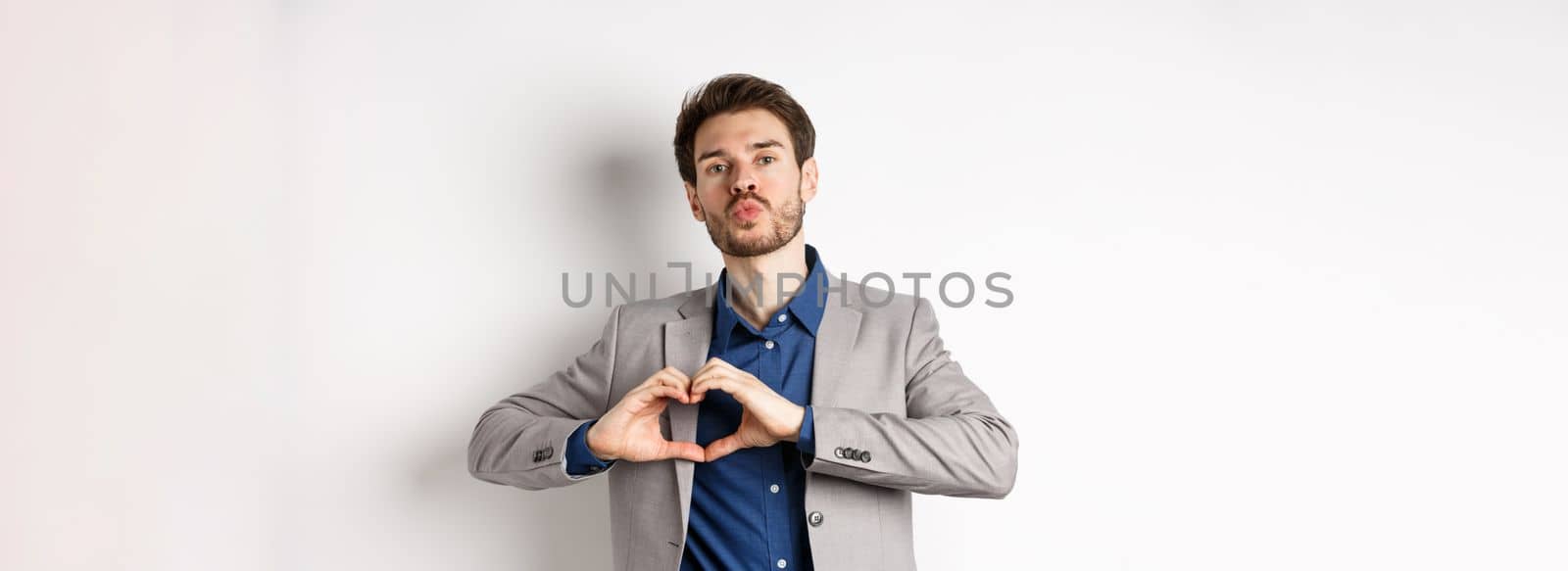 Romantic man in suit showing heart sign and pucker lips for kiss, express love and passion, like his lover, standing on white background.