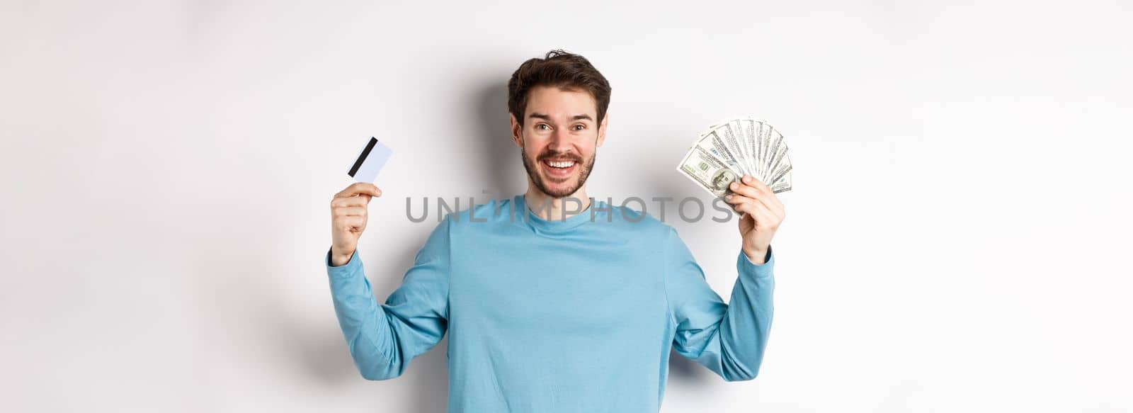 Happy guy smiling and showing plastic credit card with cash, choosing between money and contactless payments, white background by Benzoix