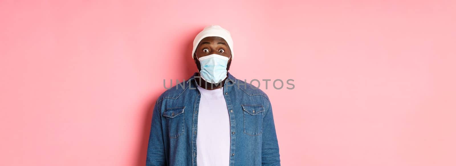 Covid-19, lifestyle and quarantine concept. Image of impressed african-american man in face mask staring at camera, gasping surprised, standing over pink background by Benzoix