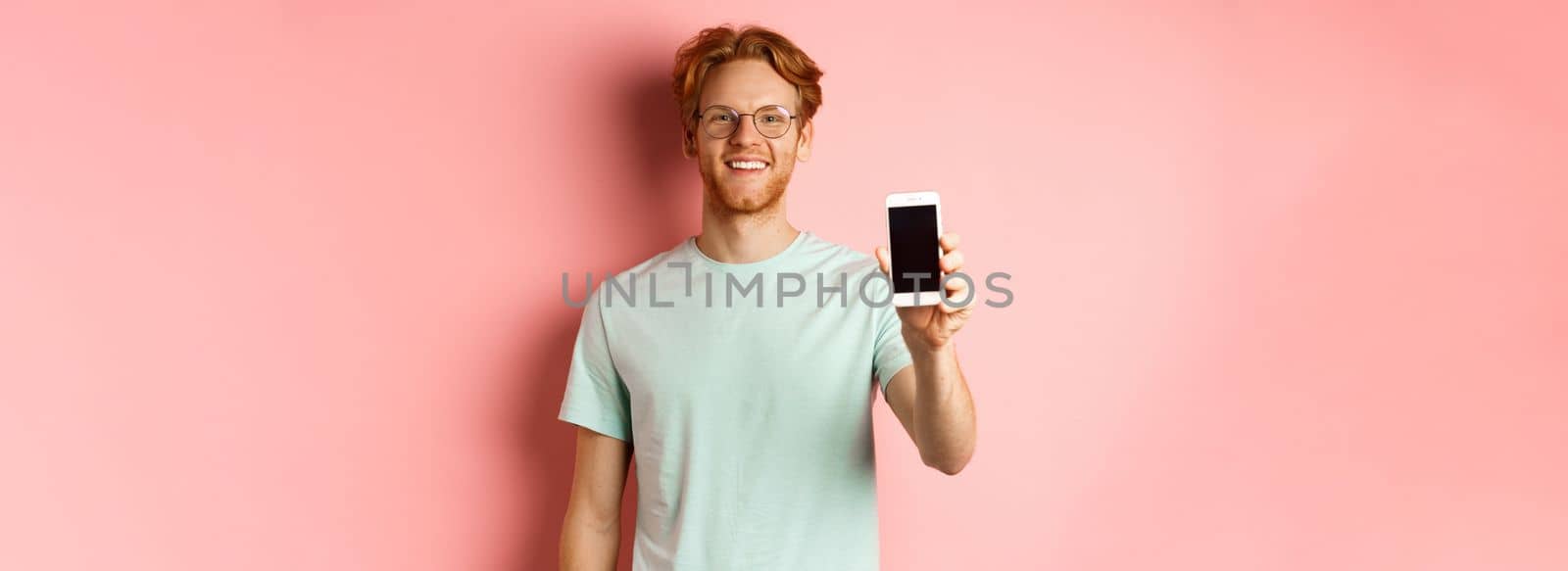 Handsome redhead man in glasses showing blank smartphone screen and smiling, demonstrate online promo or application, standing over pink background.