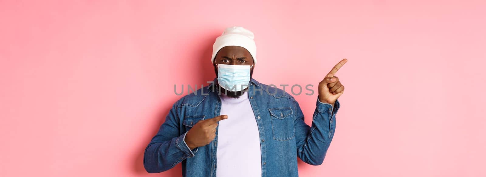 Coronavirus, lifestyle and global pandemic concept. Angry and disappointed african-american man in face mask pointing left, staring at camera displeased, pink background.
