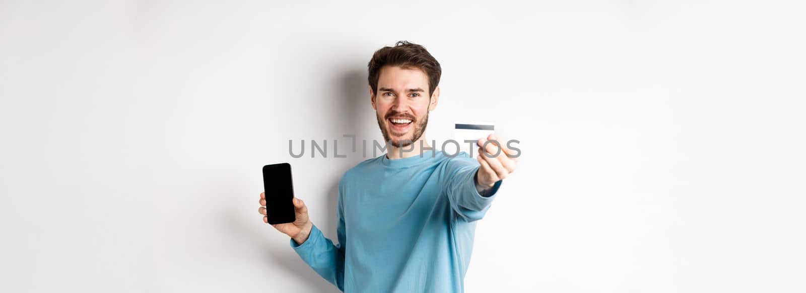 E-commerce and shopping concept. Happy man showing plastic credit card and screen of smartphone, recommending banking app, standing on white background.