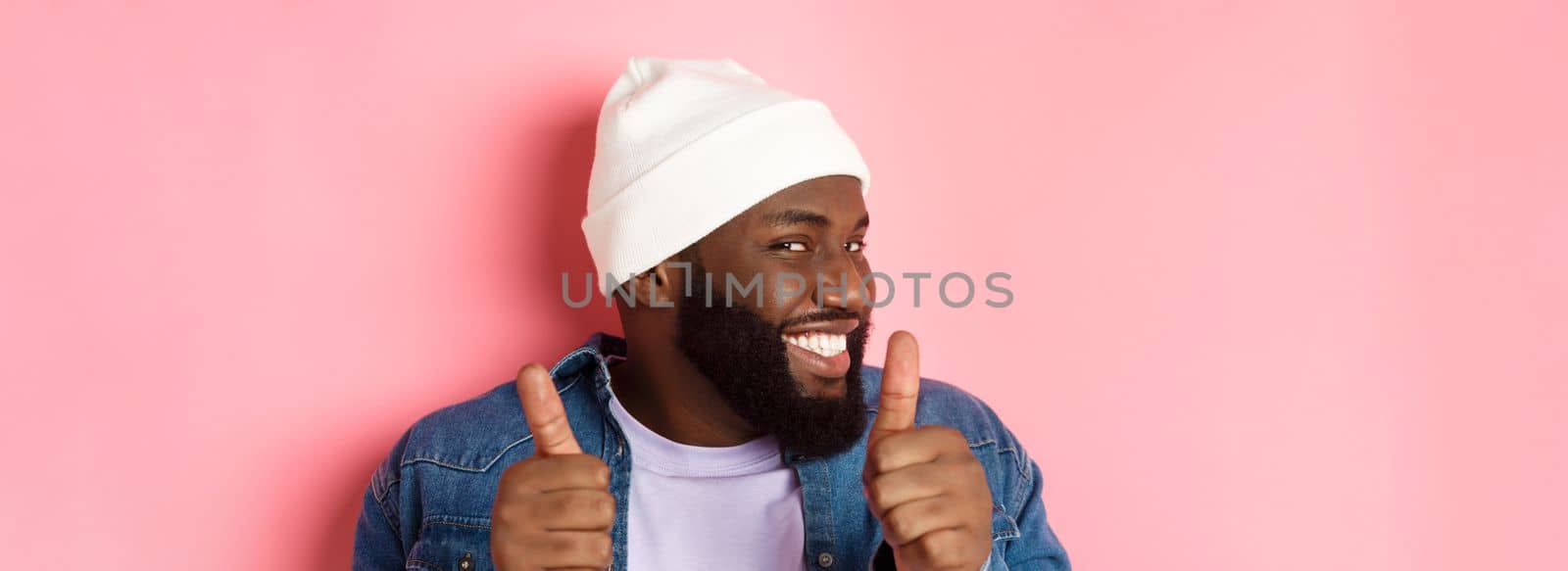 Close-up of happy Black bearded guy in beanie showing support, agree or approve something, giggle devious and showing thumbs-up, standing over pink background.