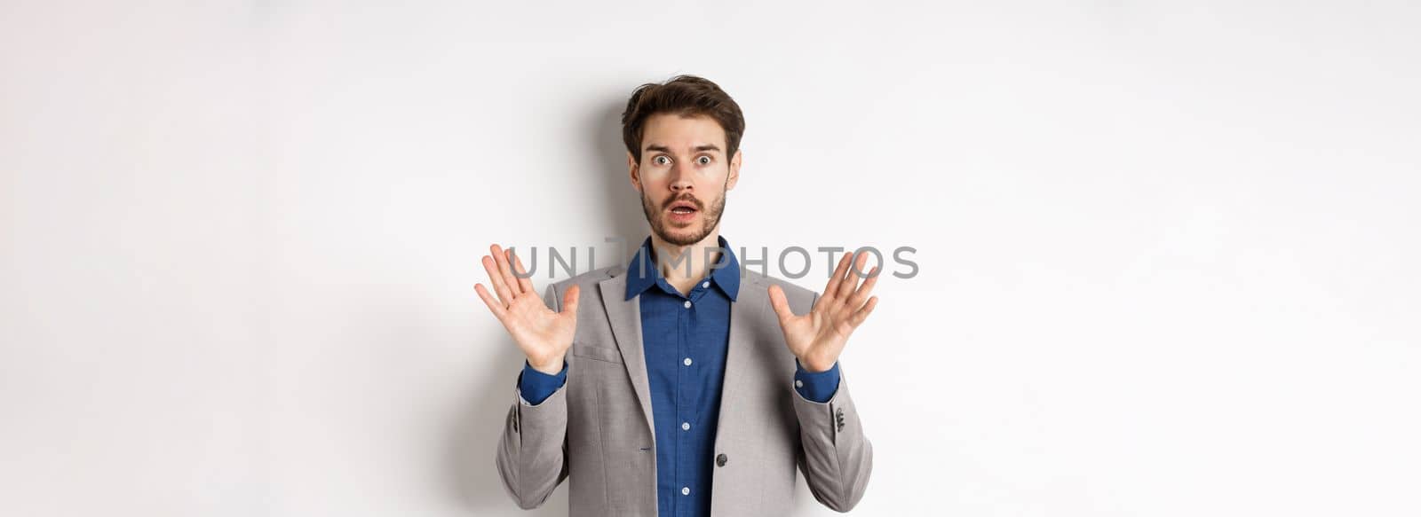Surprised businessman in suit raising hands up and look excited, standing against white background.