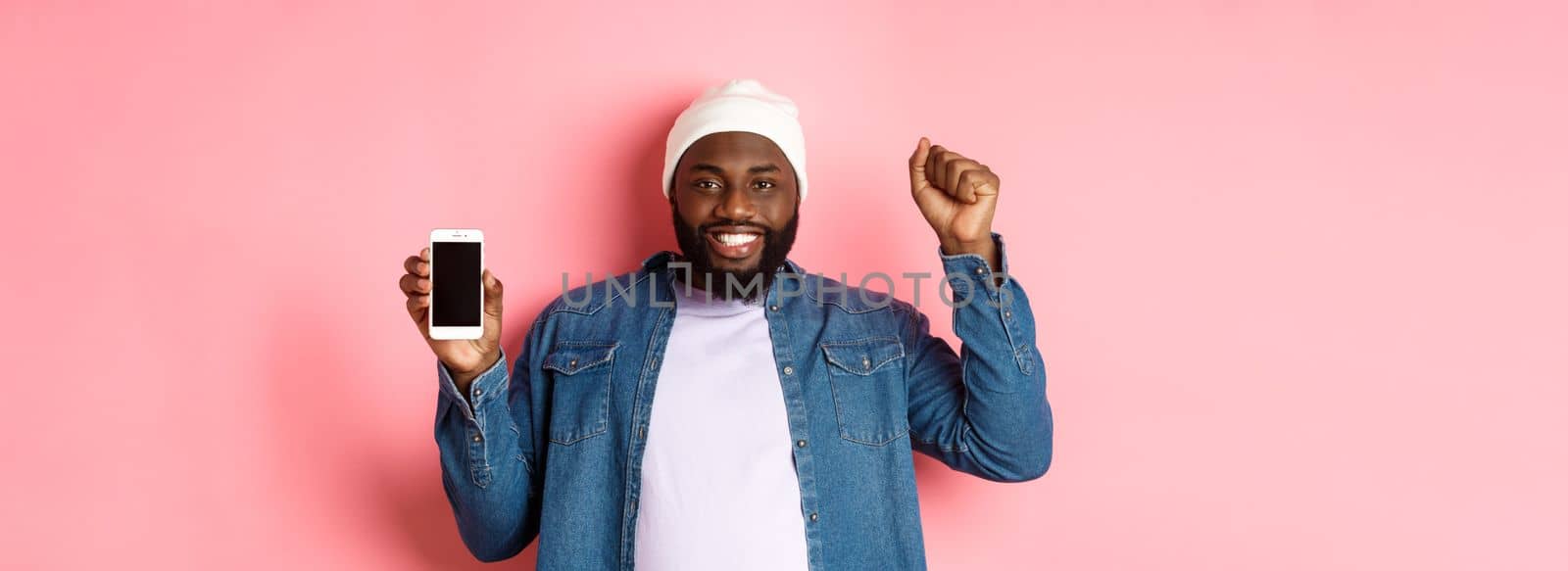 Online shopping and technology concept. Cheerful Black man rejoicing and showing mobile screen, raising hand up satisfied, triumphing while standing over pink background.