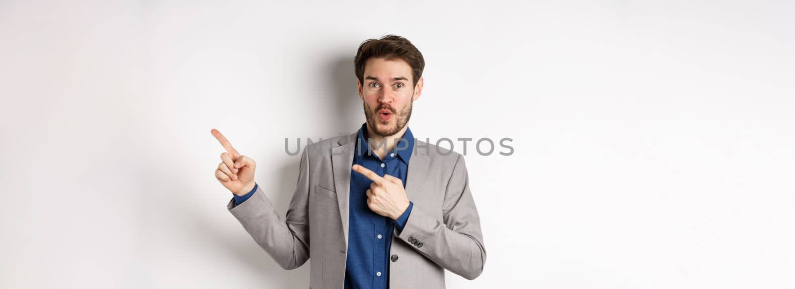 Excited handsome caucasian businessman in suit pointing right at logo, say wow and look amazed, standing on white background.