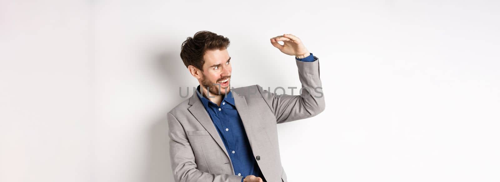 Smiling happy businessman in suit showing large size object, shaping big thing with hands and look pleased, standing on white background.