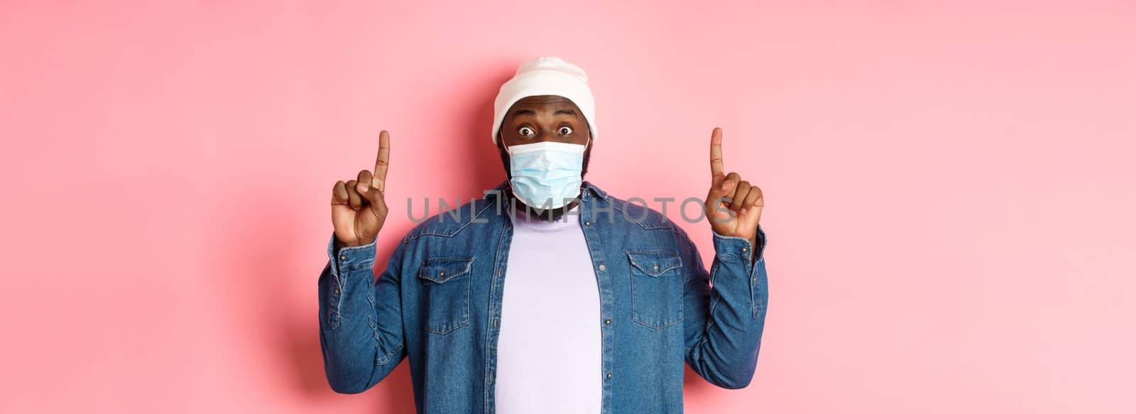 Coronavirus, lifestyle and global pandemic concept. Excited african-american guy in medical mask showing announcement, pointing fingers up, standing over pink background.