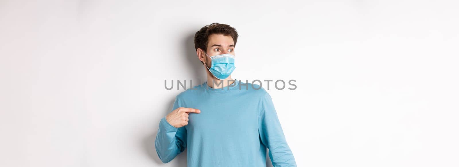 Covid-19, health and quarantine concept. Confused man in medical mask pointing at himself, looking left with ambushed face, standing over white background.