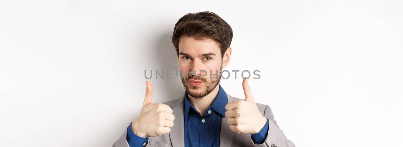 Well done. Satisfied businessman in suit showing thumbs up in approval, like and agree, recommend good company, white background by Benzoix