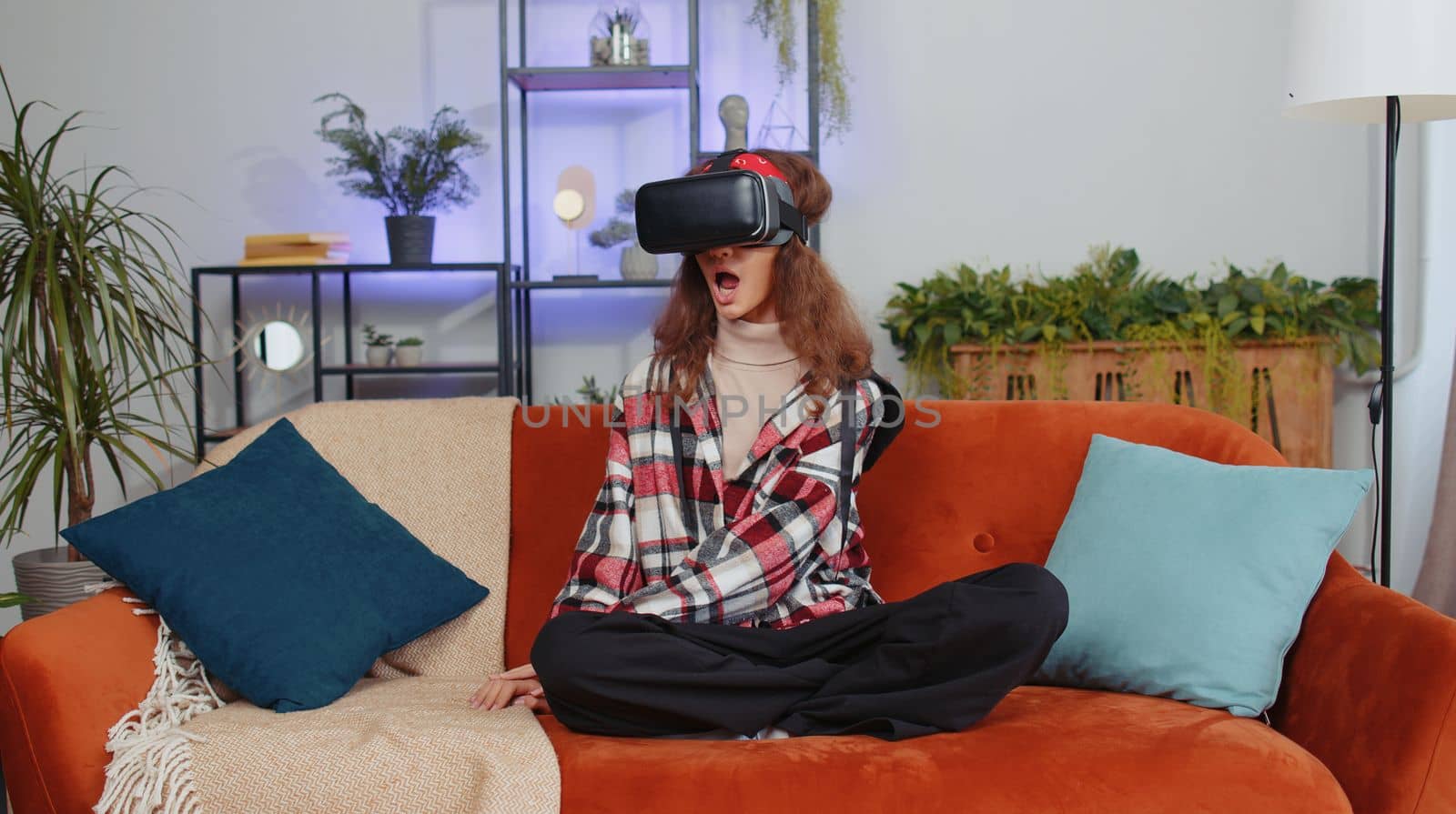 Preteen girl using virtual reality futuristic technology VR app headset helmet to play simulation 3D 360 video game, watching film movie at modern home apartment. Child kid in goggles sits on sofa