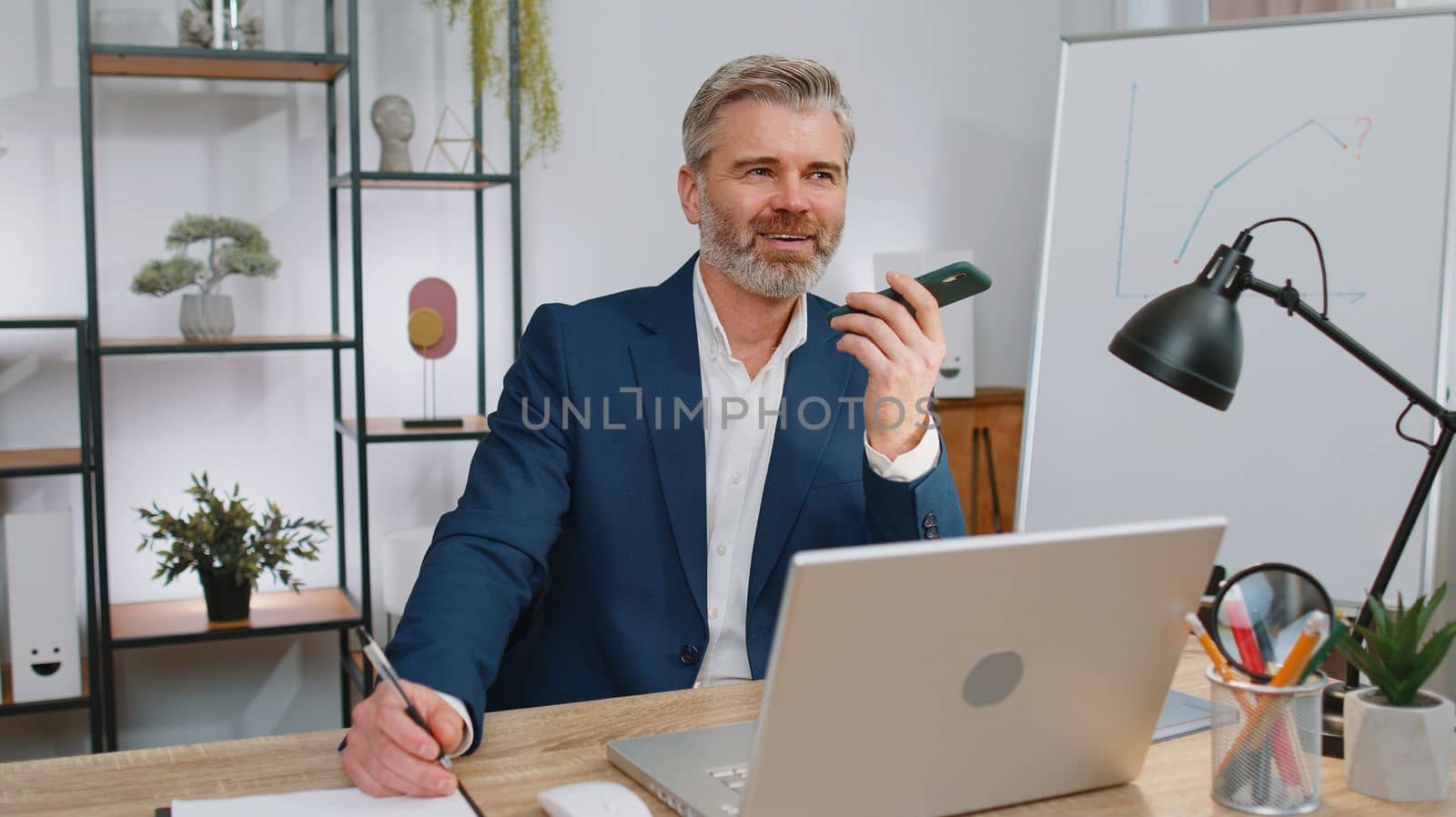 Mature businessman working, having mobile phone loudspeaker talk at office desk with laptop. Manager freelance old gray-haired man holding smartphone using messenger chat apps. Employment, occupation