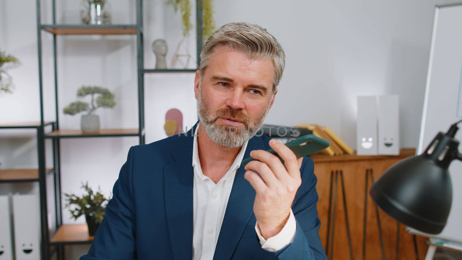 Mature businessman working, having mobile phone loudspeaker talk at office desk with laptop. Middle aged professional freelance man holding smartphone using messenger chat apps. Employment, occupation