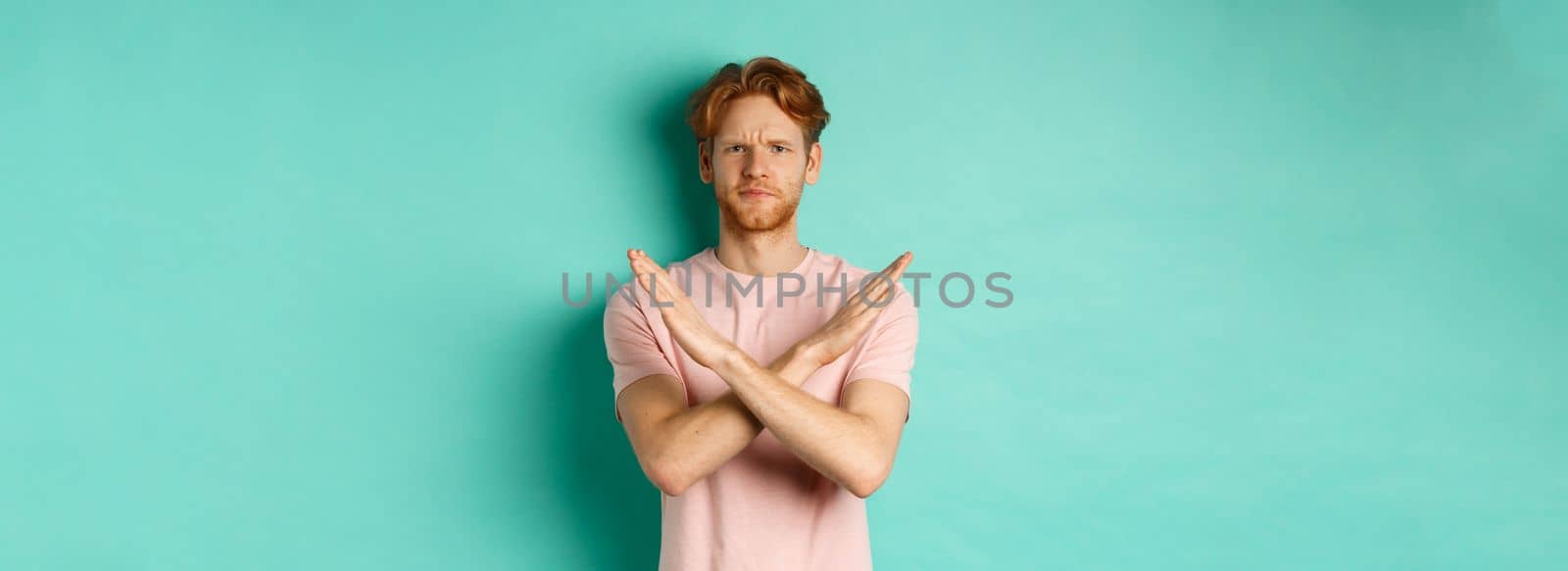 Skeptical redhead man in t-shirt saying no, cross arms on chest and frowning disappointed, prohibit something bad, standing over turquoise background.