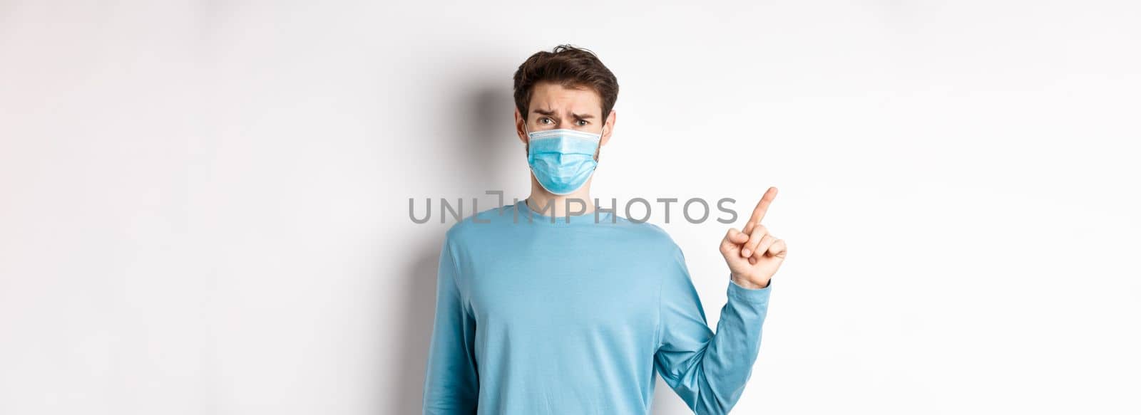 Coronavirus, health and quarantine concept. Skeptical man in medical mask pointing finger left at logo, frowning disappointed, standing over white background.