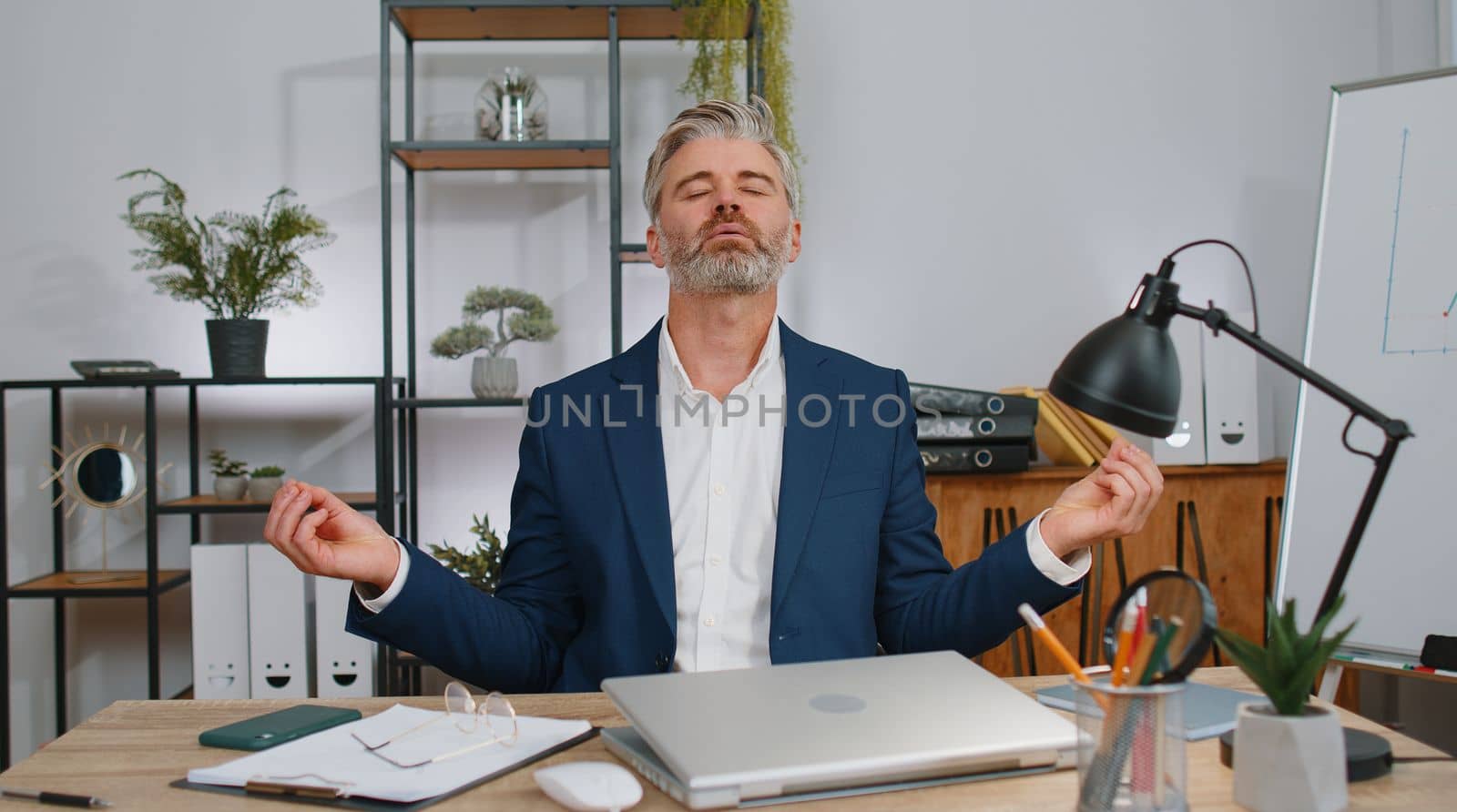 Mature businessman working on laptop computer, meditating, doing yoga breathing exercise in lotus position at home office. Calm serene freelancer old man taking break. Busy occupation. Peace of mind