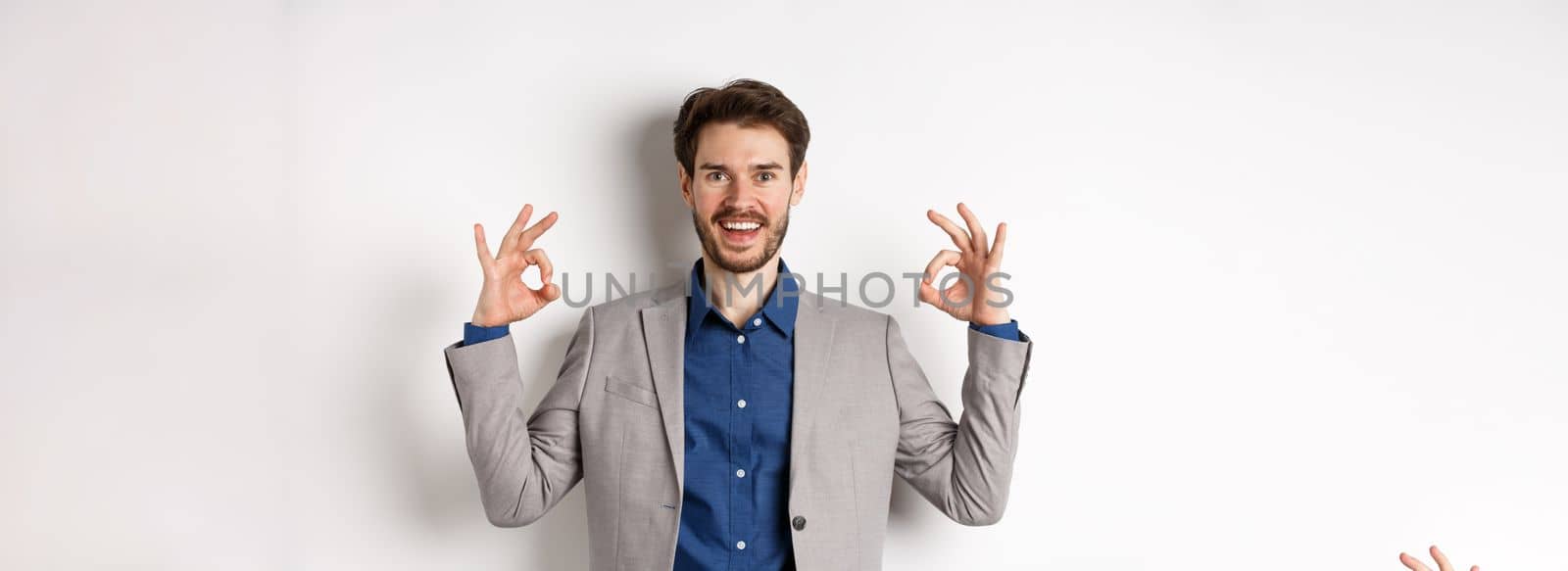 Good choice. Smiling businessman in suit showing okay signs and look happy, recommending good deal, like and approve your choice, standing satisfied on white background.