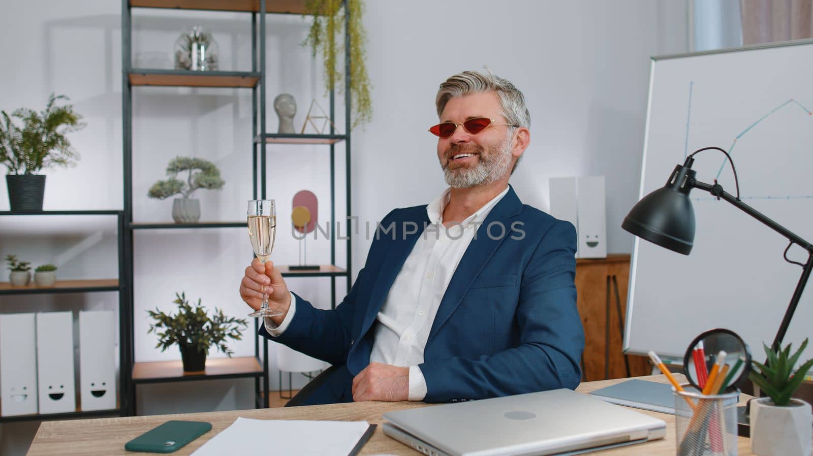 Mature old business man wears sunglasses, festive cap, drinking champagne at workplace dancing victory dance, weekend. Rejoicing developing project. Celebrating success. Lottery jackpot. Office people