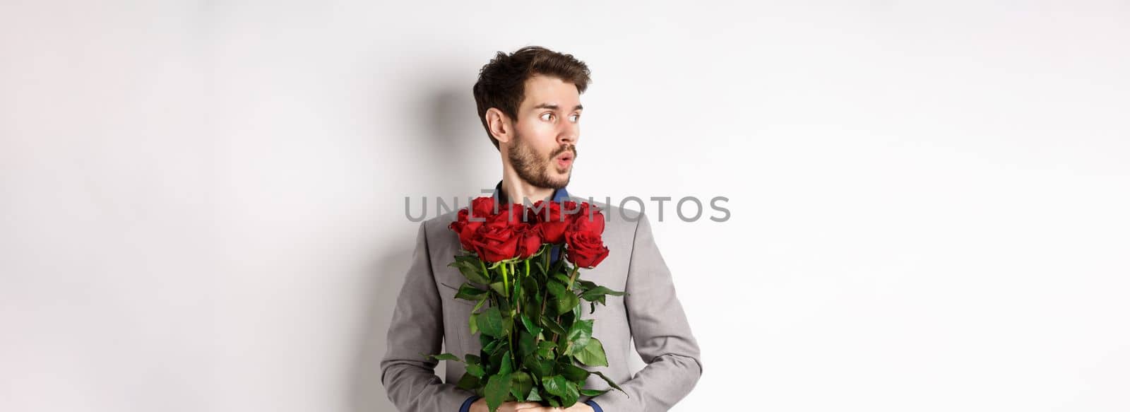 Nervous boyfriend waiting for his date on valentines day, looking left amazed, holding bouquet of red roses, standing in suit over white background.