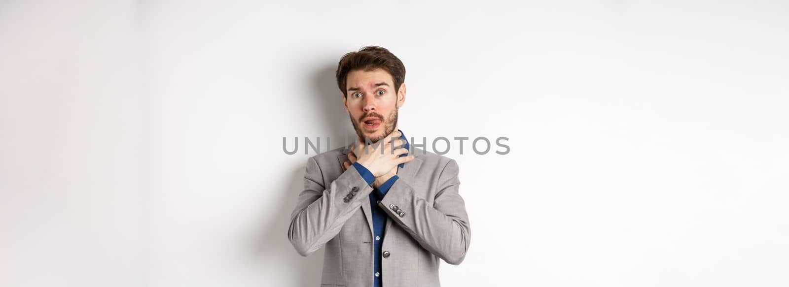 Distressed young man in business suit strangle himself and showing tongue, feeling sick with sore throat, standing on white background.