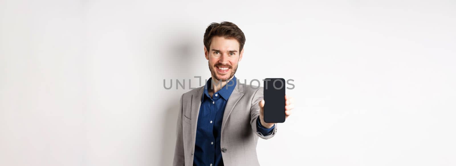 E-commerce and online shopping concept. Confident businessman in suit stretch out hand with empty smartphone screen, showing on phone, standing against white background.