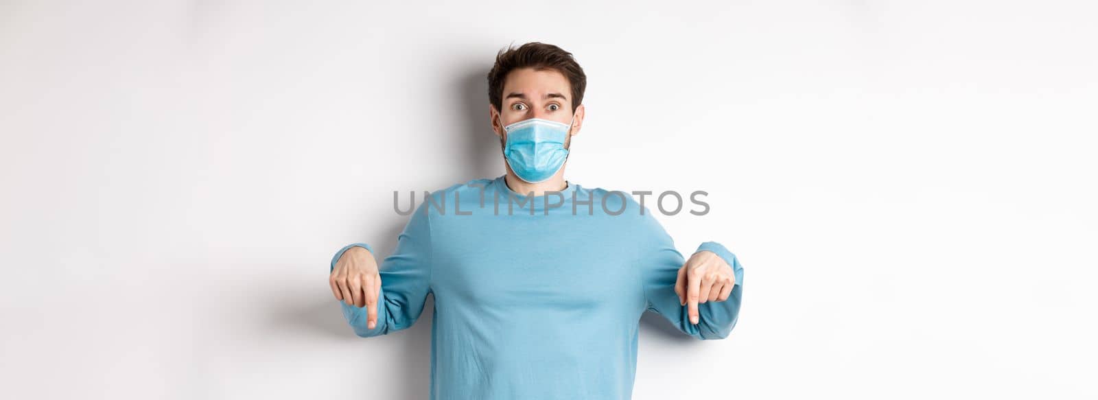 Covid-19, health and quarantine concept. Amazed man checking out promo logo, pointing fingers down, wearing medical mask from coronavirus, white background.