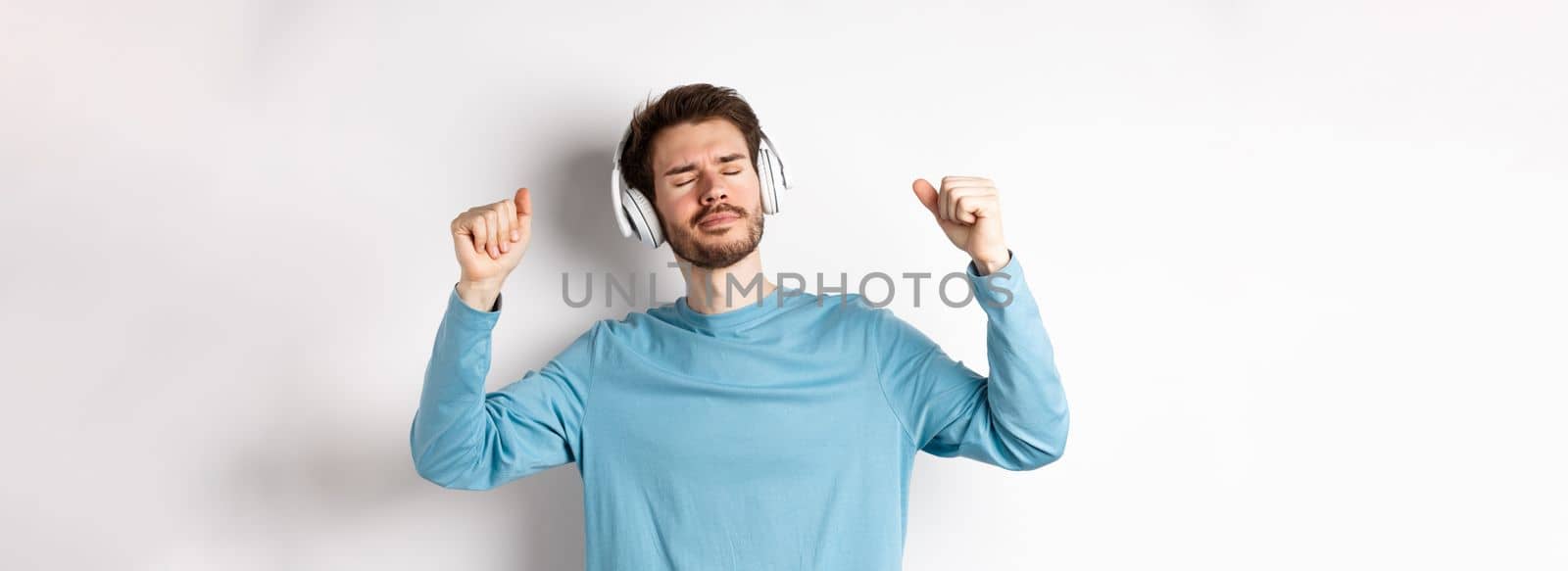 Happy handsome man dancing to music in headphones, listening songs and smiling, standing over white background.
