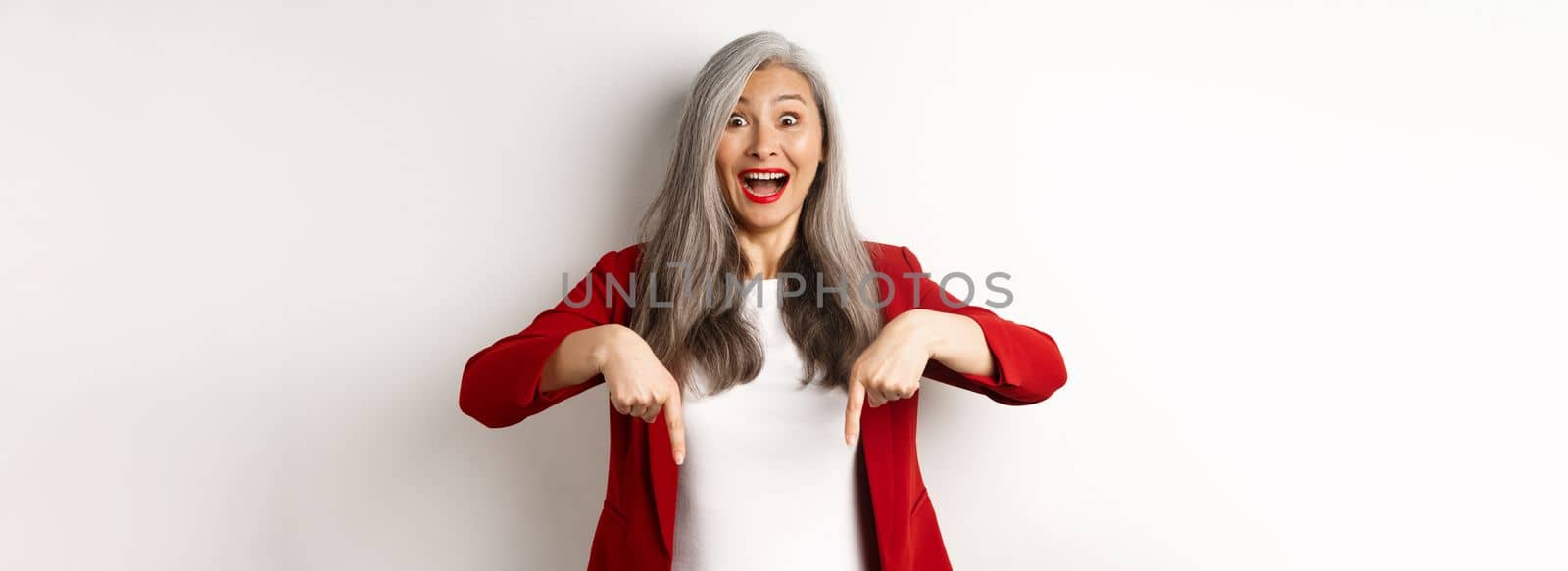 Portrait of happy asian lady in red blazer showing logo, pointing fingers down and smiling cheerful, check this out gesture, white background.