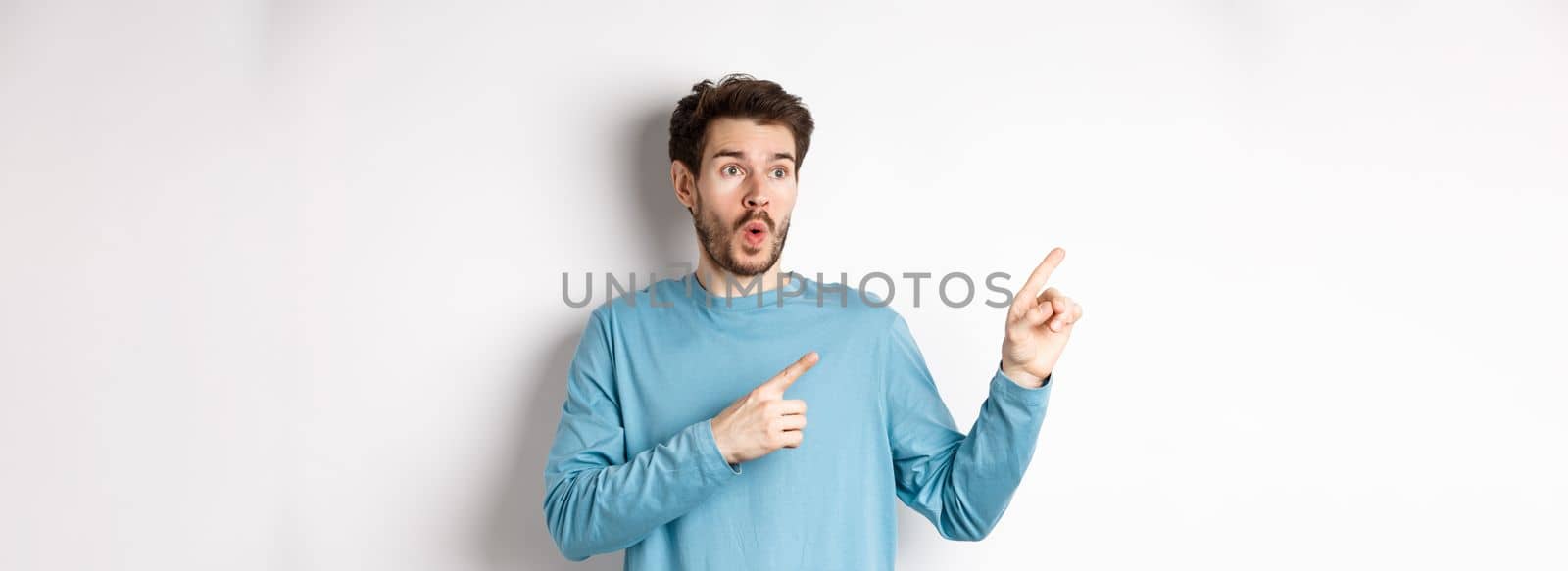 Wow check it out. Impressed young man pointing and looking left at special promo offer or discounts on white background, gasping amazed, standing in studio by Benzoix