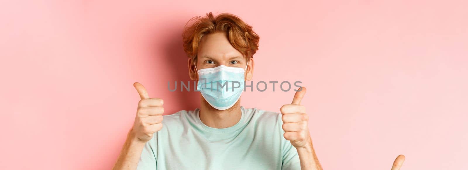 Covid-19 and pandemic concept. Handsome guy with messy ginger hair, wearing medical mask on face and showing thumbs up, standing over pink background.