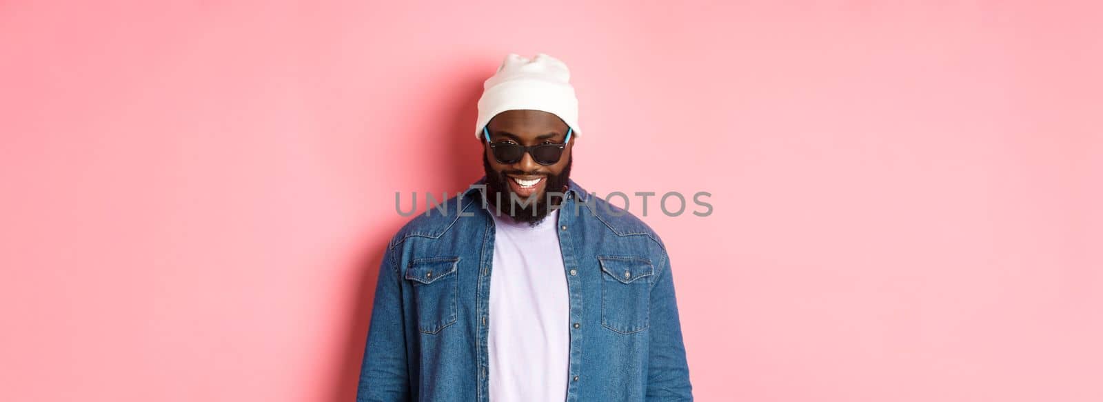 Cool african-american hipster man in beanie and sunglasses, smiling pleased and looking at camera, standing confident over pink background.