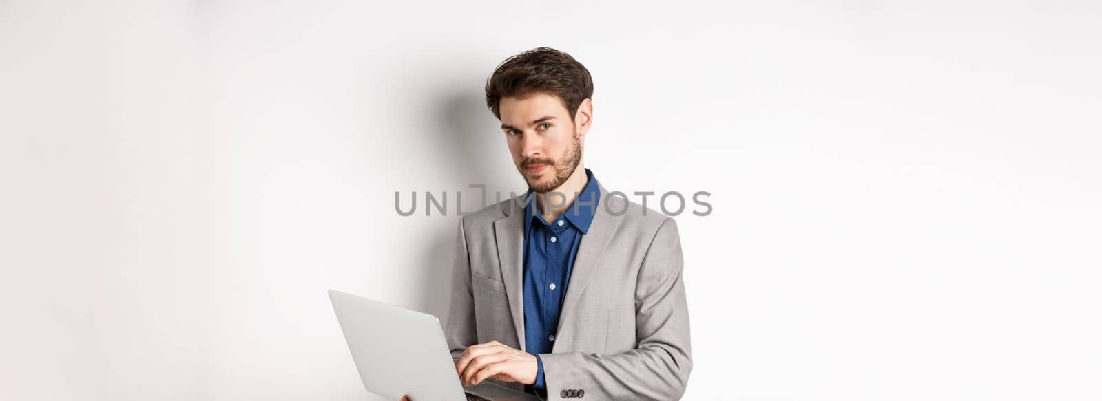 Handsome businessman in suit working on laptop, looking at camera confident, standing against white background.