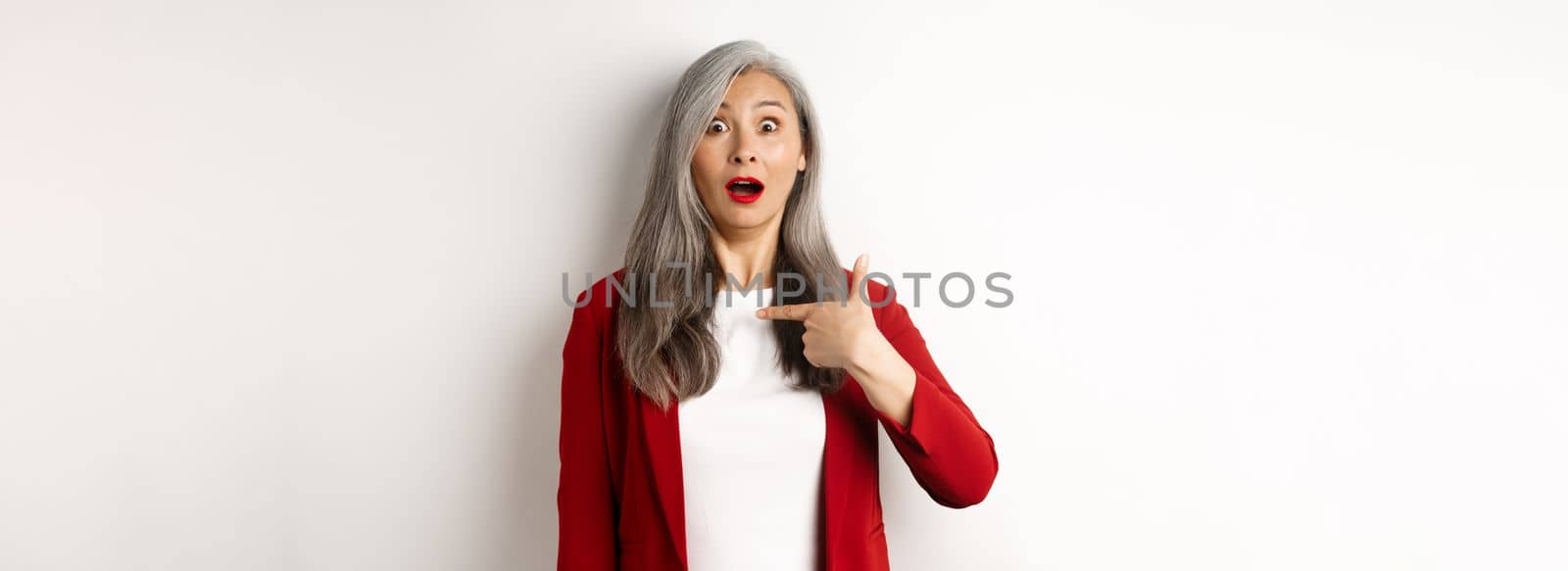 Surprised asian woman with grey hair, pointing at herself and gasping confused, standing over white background.