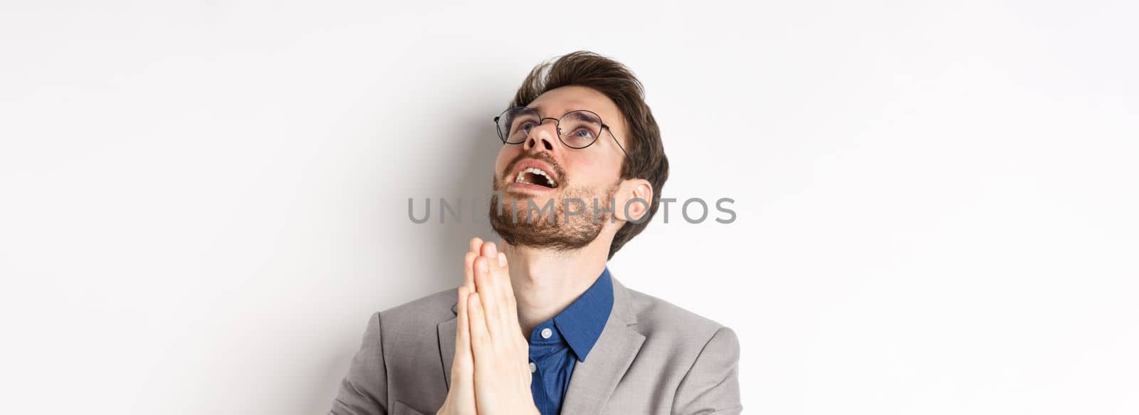 Close-up of handsome bearded man in glasses and suit looking up and praying god, begging lord for something, standing on white background.