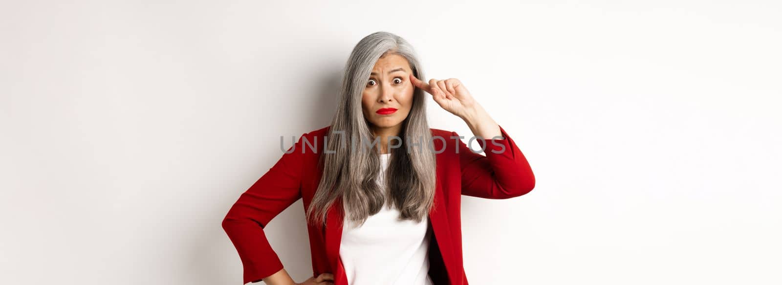 Annoyed asian business woman scolding someone stupid, showing crazy gesture, pointing finger at head and staring at camera, white background.