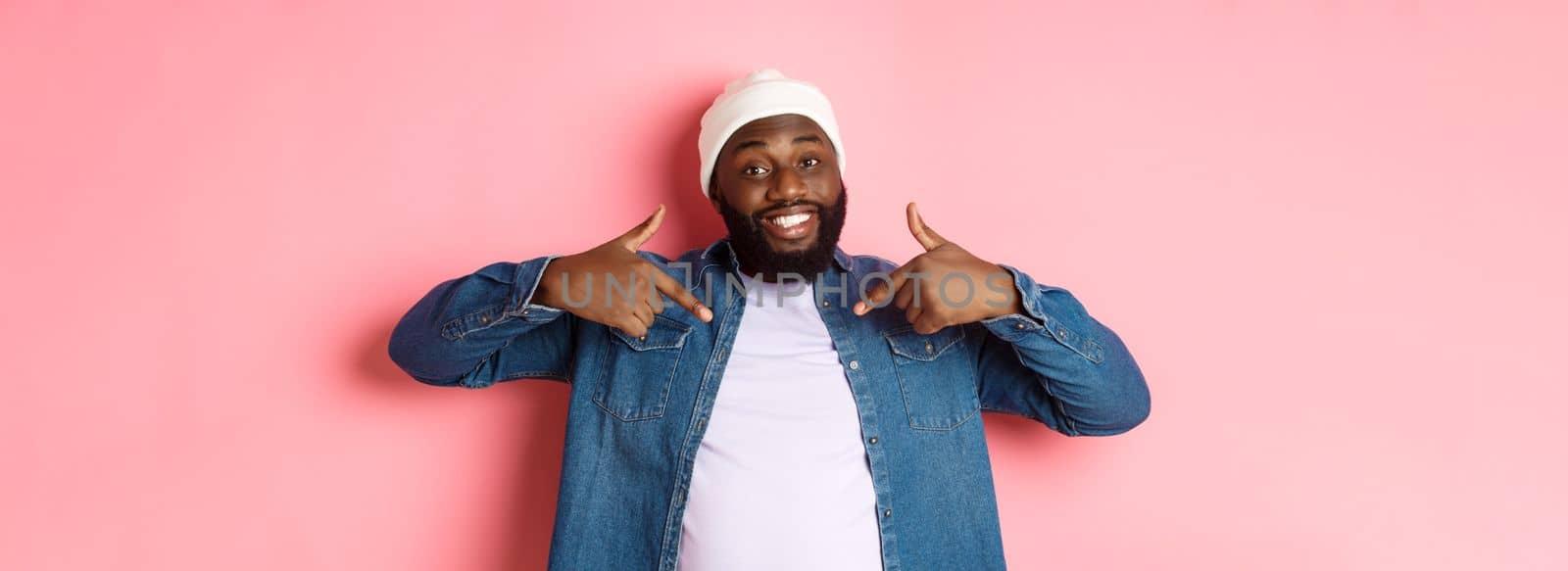 Happy young african-american hipster guy pointing at himself t-shirt, smiling pleased, standing over pink background.