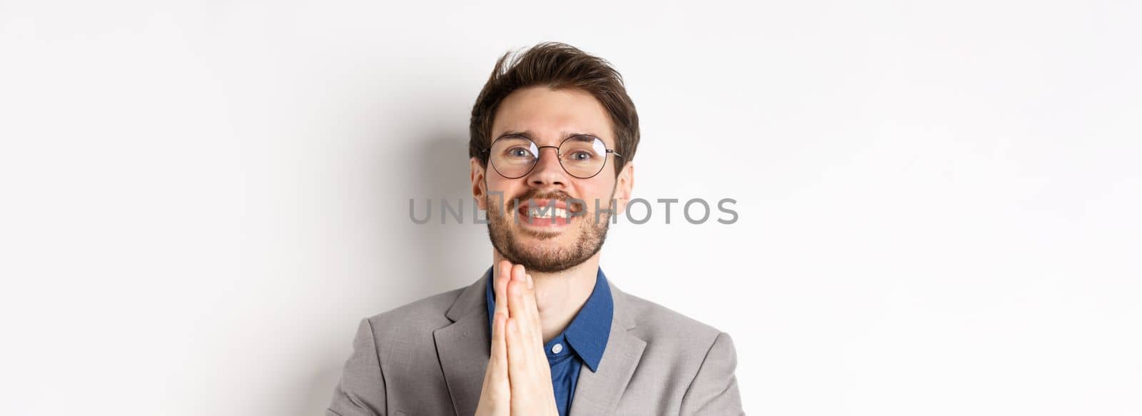 Close-up of happy thankful guy in glasses and suit showing gratitude, make namaste sign and smiling, standing on white background.