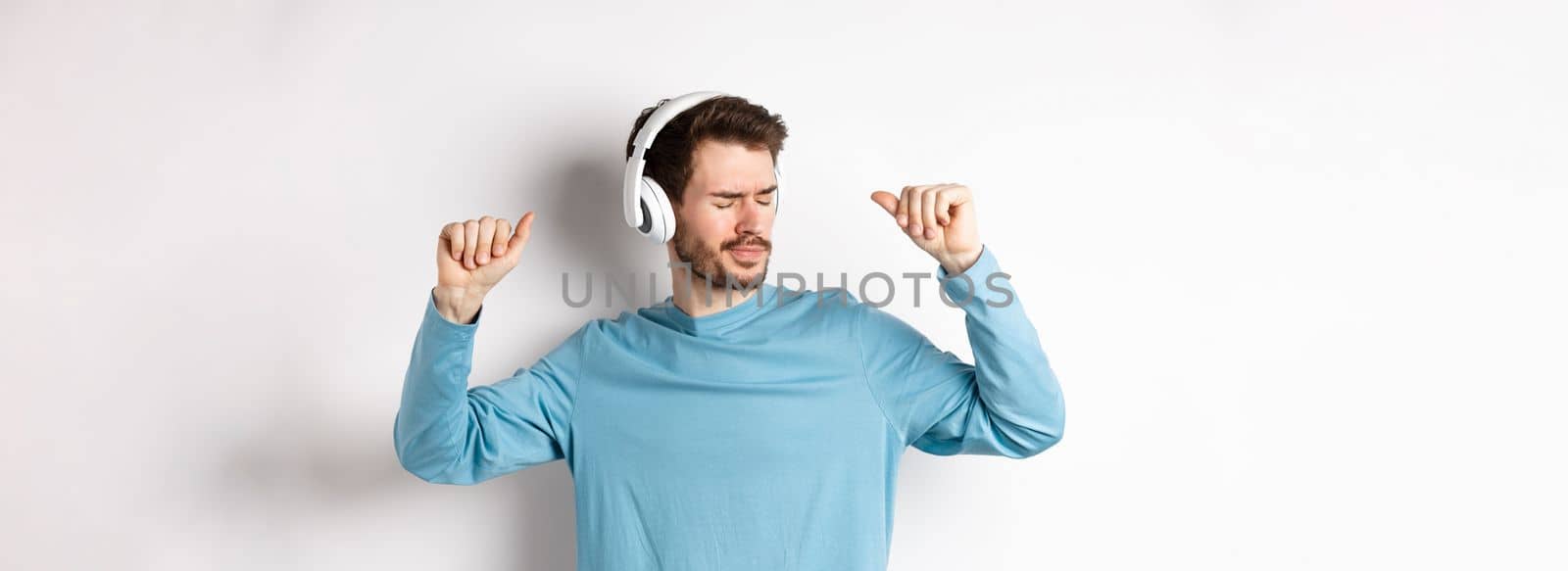 Happy young man having fun in headphones, dancing while listening music in wireless earphones, white background.
