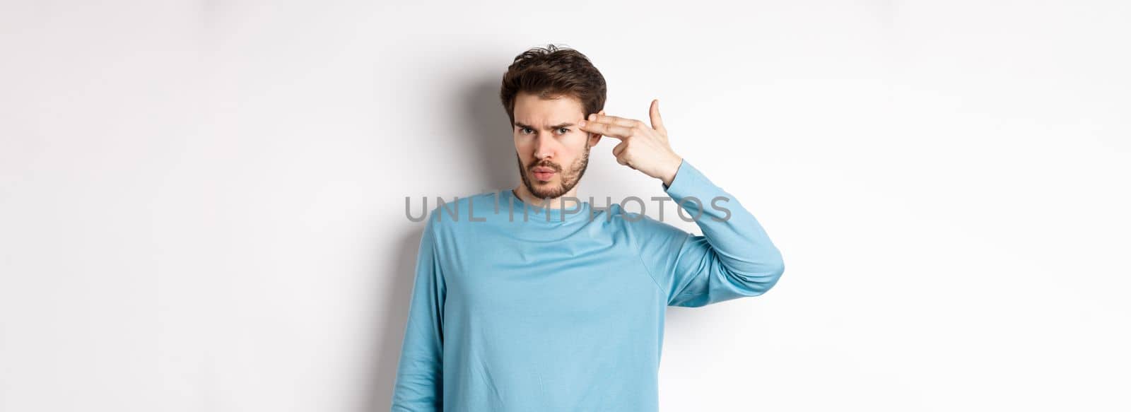 Annoyed young man frowning, showing shooting gun near head gesture, blow brain out sign, standing over white background.