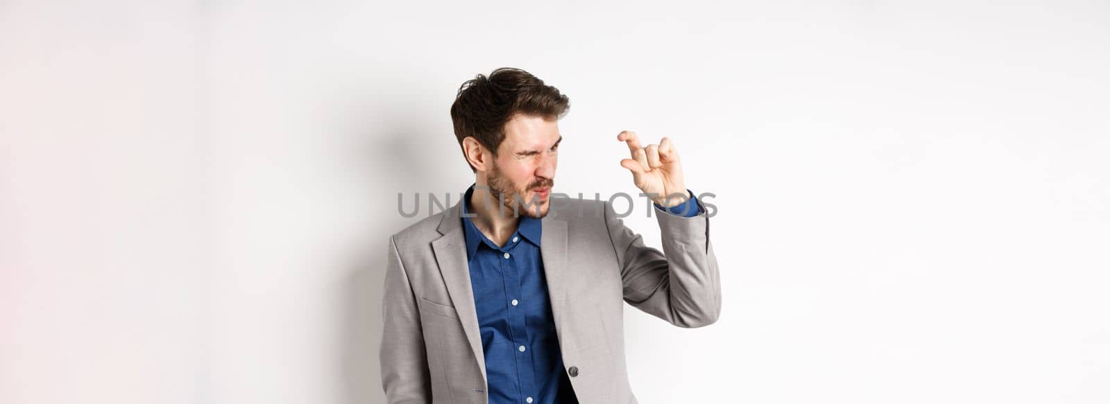 Businessman trying to see something tiny, showing little thing size with fingers, standing in suit on white background.