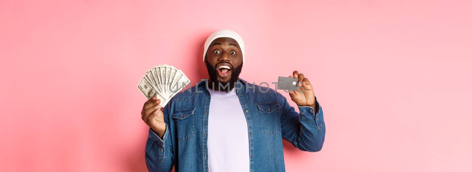 Shopping concept. Excited african-american man showing credit card and dollars, got deposit or money loan, standing over pink background.