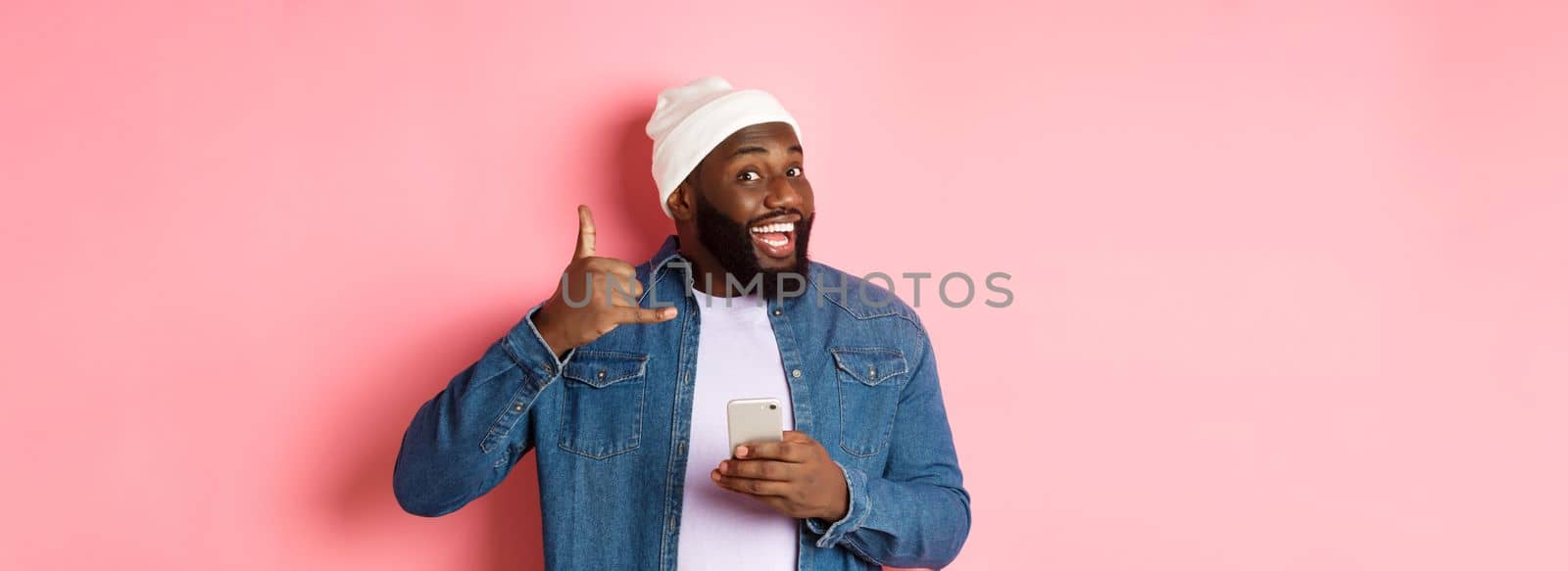Happy Black man showing call me sign, making phone gesture and smiling, holding smartphone, standing in beanie and denim shirt over pink background.