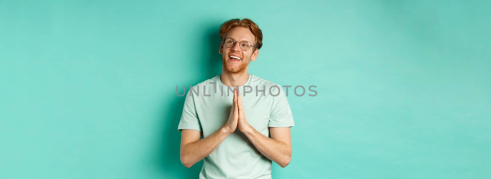 Hopeful redhead man with beard, wearing glasses and t-shirt, holding hands in namaste or plead gesture and looking right, smiling and thanking, standing over turquoise background.