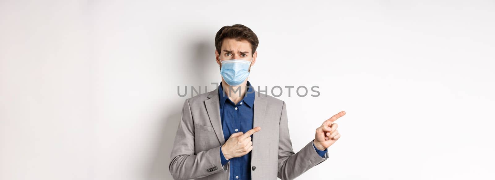 Covid-19, pandemic and business concept. Upset man in medical mask and office suit, frowning sad and pointing left at banner, standing on white background.