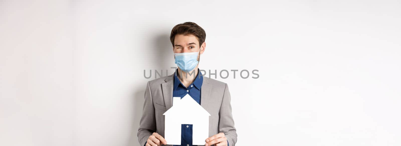 Real estate and covid-19 concept. Cheerful young man in medical mask and suit winking at camera, advertising new flats, showing paper house cutout.
