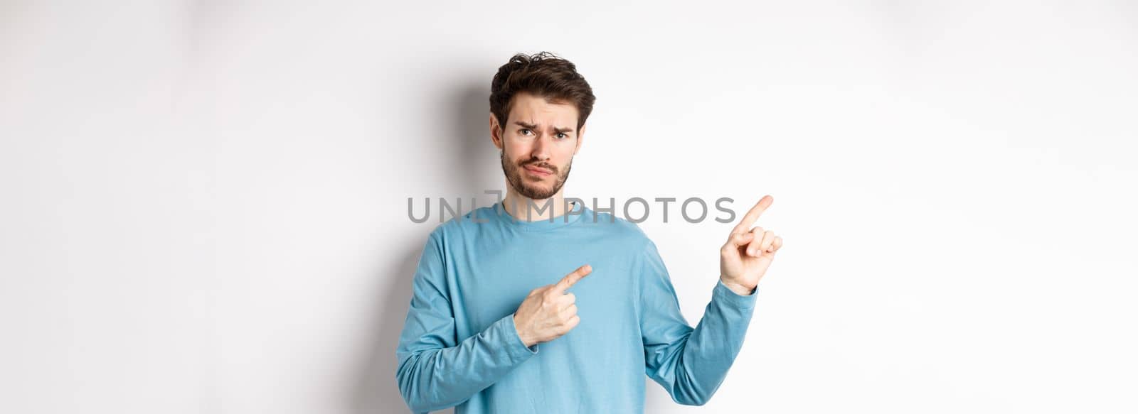 Skeptical young man with beard grimacing unsatisfied, frowning and pointing at upper left corner logo doubtful, standing upset over white background.